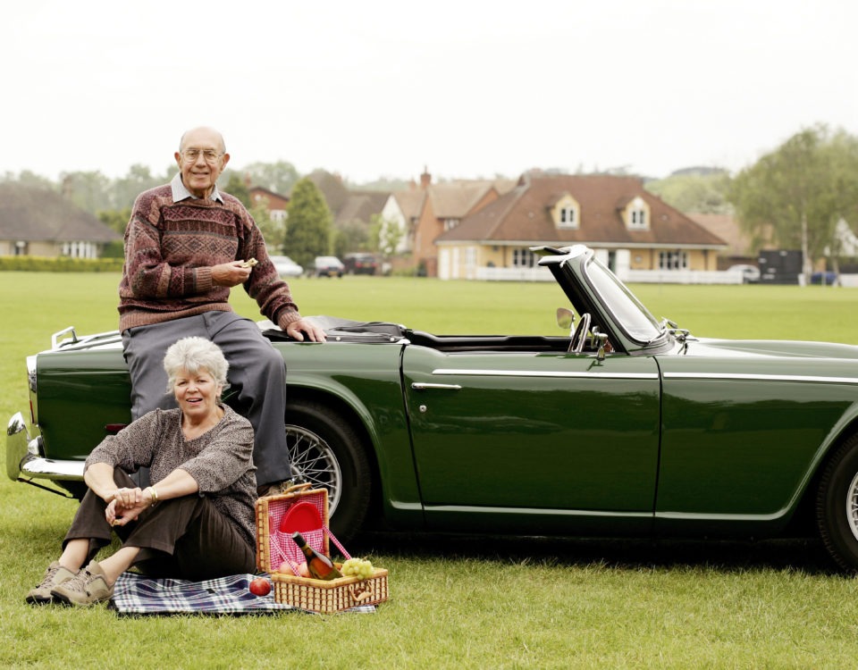 Ein älteres Ehepaar macht ein Picknick auf einer grünen Wiese neben Ihrem Oldtimer.